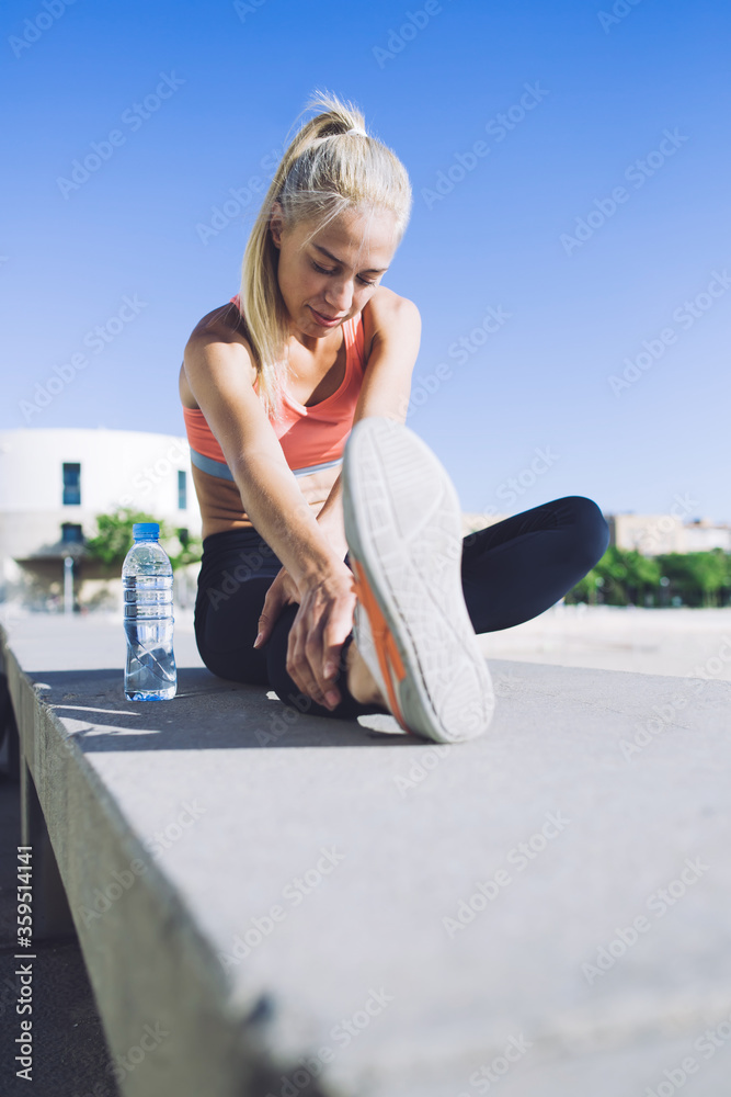 Portrait of a beautiful athletic female stretching legs muscles while sitting on concrete pter in summe summer day, young fit woman doing warm up exercises before begin morning run in the fresh air