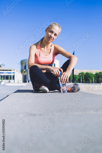 Portrait of a smiling fit woman sitting with bottle of water while taking break after fitness training in the fresh air in sunny summer day, young female runner resting after workout outdoors © BullRun