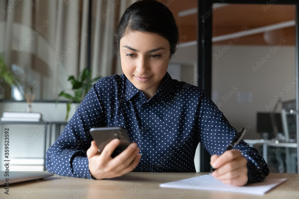 Indian businesswoman sit at desk hold cell look at screen distracted for writing report check sms read received message got reminder. Student do task use internet web search helpful info prepare essay Indian businesswoman sit at desk hold cell look at screen distracted for writing report check sms read received message got reminder. Student do task use internet web search helpful info prepare essay