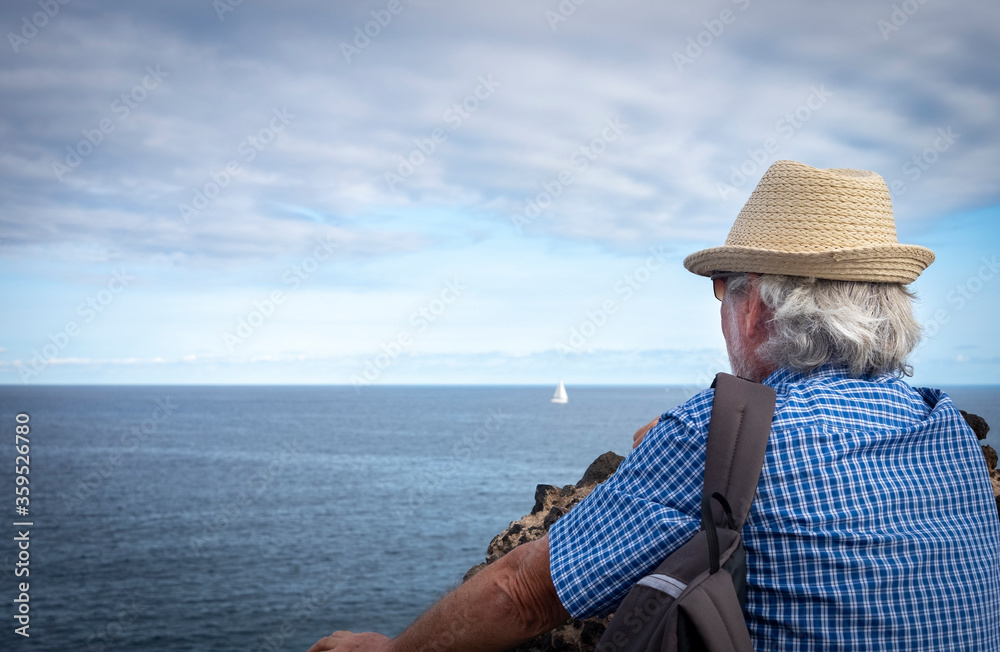 Shoulders view of senior adult man with straw hat in sea excursion with backpack looking at the blue ocean.  Happy and free active retirement concept for an elderly people