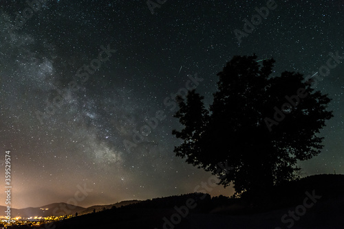 Sternenhimmel mit der Milchstraßen Galaxie im Sommer mit einem Baum vor einer beleuchteten Stadt, Deutschland