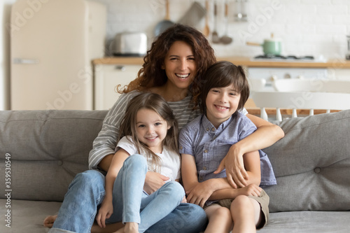Close up portrait picture of happy young mother with children sitting on couch at home. Smiling parents with little kids hugging looking at camera posing for photo in living room.