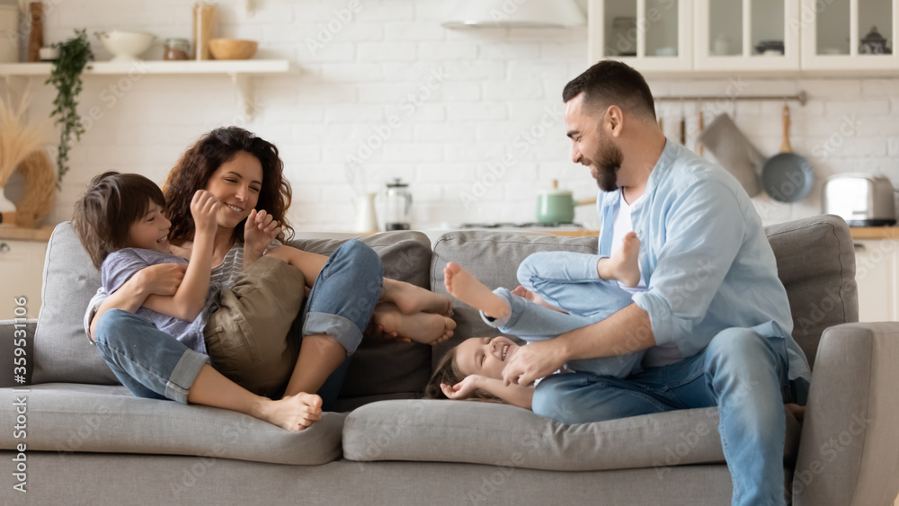 Happy father and mother tickling young daughter and son sitting on couch in living room. Family having fun at home. Parents playing with cute daughter and son Happy father and mother tickling young daughter and son sitting on couch in living room. Family having fun at home. Parents playing with cute daughter and son