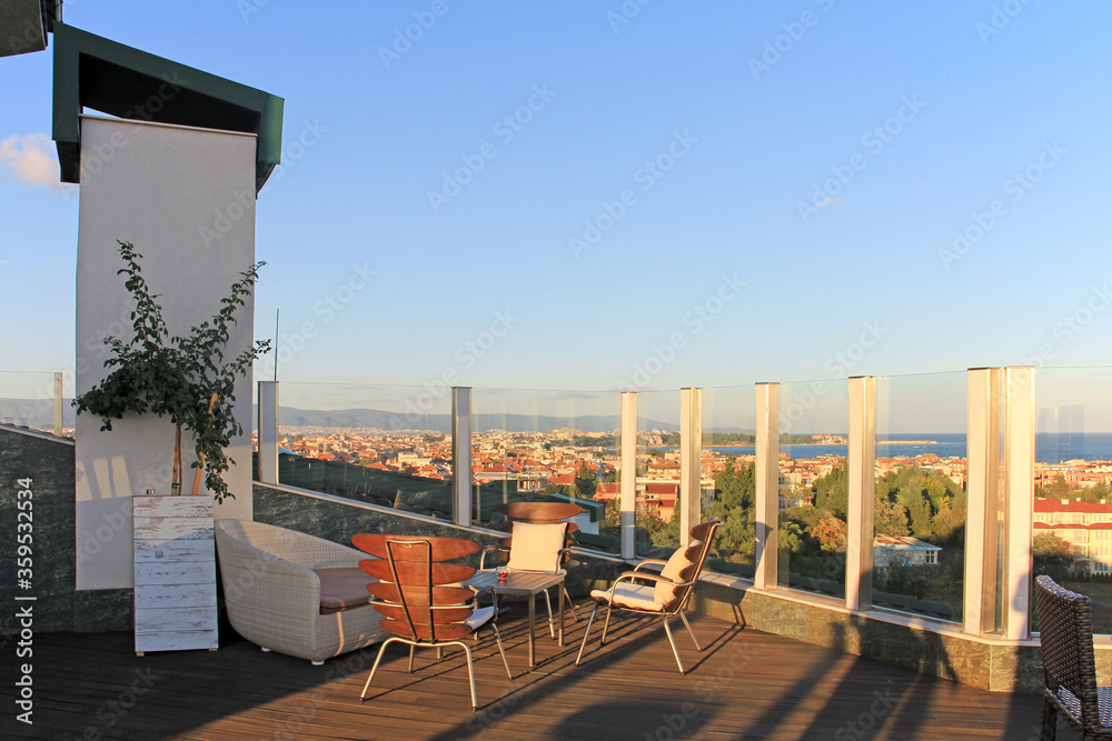 Outdoor cafe on the terrace with sea views. Recreation area with modern furniture: sofa, table and chairs on a high roof with glass railings. Panoramic evening summer view of the city by the sea.