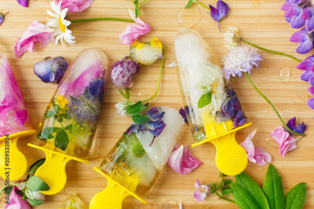 Fototapeta premium Hello summer concept. Floral Ice Pops flat lay. Colorful wildflowers in frozen popsicles and ice cubes and fresh summer flowers on wooden background. Creative image