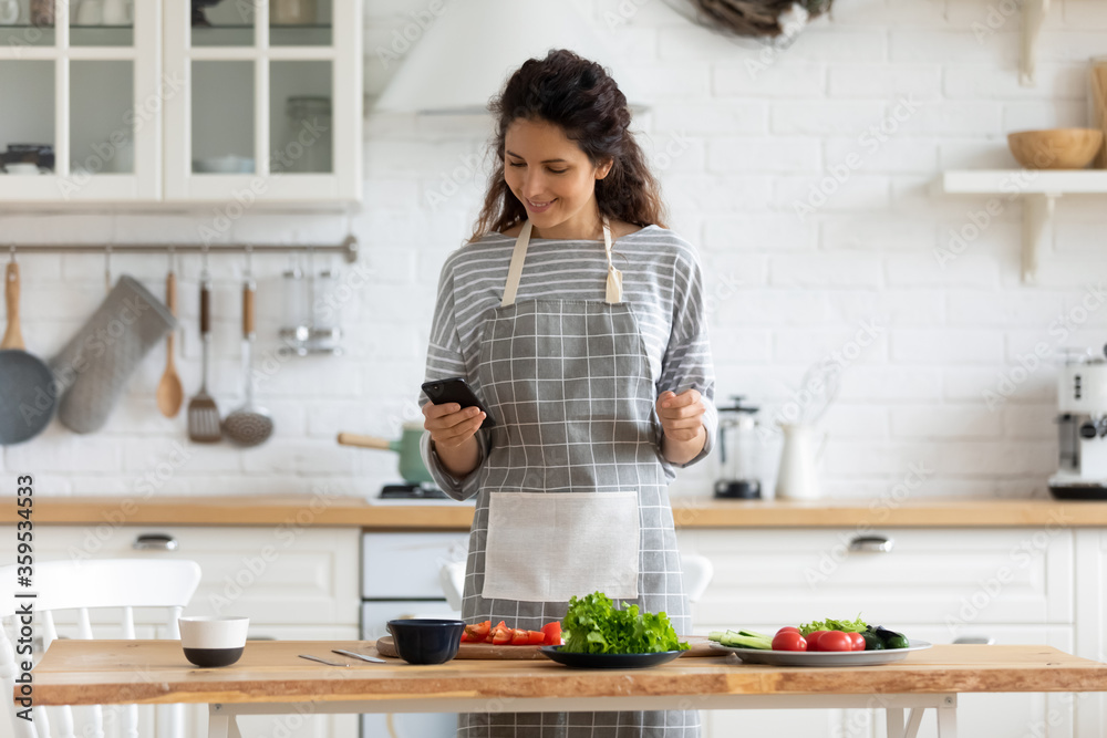 Happy young attractive woman cooking to recipe on smartphone in kitchen at home. Smiling woman in apron taking online web courses for preparation dinner with organic fresh meal.