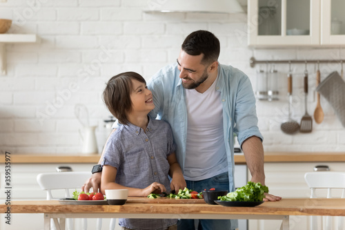 Caring father teaching little son top use knife looking at each other standing at table in kitchen. Happy dad and adorable boy chopping vegetables lettuce on wooden cutting board, cooking salad.