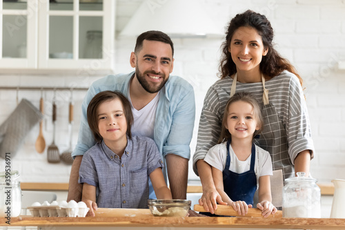 Close up headshot portrait picture of happy young couple with children cooking dinner looking at camera standing at table in kitchen. Smiling family using rolling pin for pastry on wooden board.