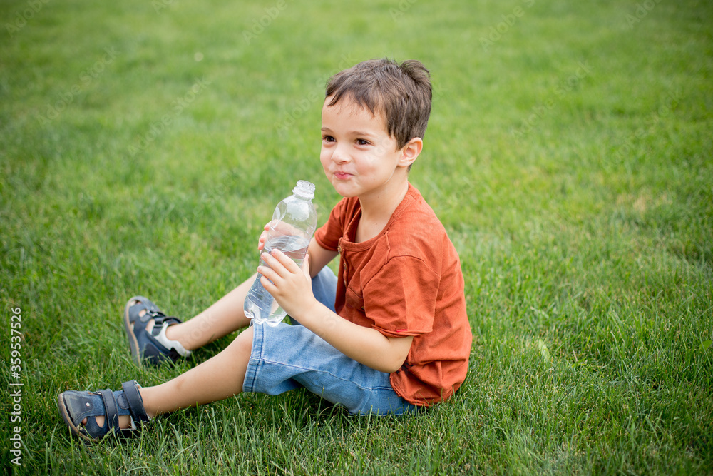 Cute cheerful child sits on a lawn with a plastic bottle of water in his hands. Bright green grass. Summer. Clothing casual. Blue jeans and a brown t-shirt. A boy of 4-6 years old is smiling. Cute cheerful child sits on a lawn with a plastic bottle of water in his hands. Bright green grass. Summer. Clothing casual. Blue jeans and a brown t-shirt. A boy of 4-6 years old is smiling.