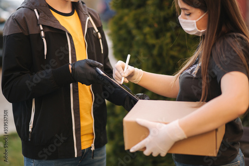 Man from delivery service in medical mask and gloves handing fresh food to young woman customer receiving express delivery from courier at home. Courier with tablet, customer sign in. Female order