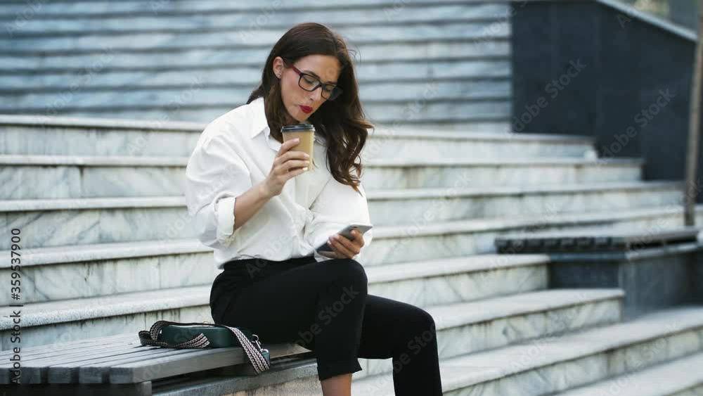 Lady in glasses, formal attire. Drinking coffee while sitting on bench stairs and using mobile phone, working online outdoors. Business concept Lady in glasses, formal attire. Drinking coffee while sitting on bench stairs and using mobile phone, working online outdoors. Business concept