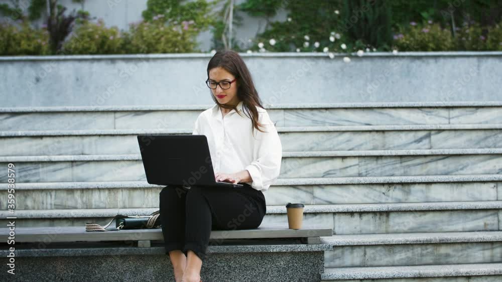 Female in glasses, white shirt and black pants. Sitting on bench stairs, typing on laptop computer, working online outdoors. Business concept Female in glasses, white shirt and black pants. Sitting on bench stairs, typing on laptop computer, working online outdoors. Business concept