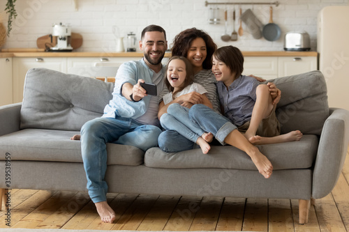 Smiling parents with little kids laughing using smartphone together sitting on couch at home. Happy father holding phone taking selfie with children. Family watching video having fun with cellphone.