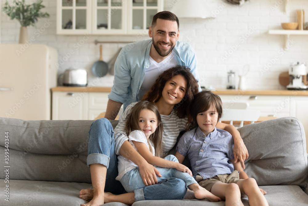 Close up portrait picture of happy young family of four people sitting on couch at home. Smiling parents with little children hugging looking at camera posing for photo in living room. Close up portrait picture of happy young family of four people sitting on couch at home. Smiling parents with little children hugging looking at camera posing for photo in living room.