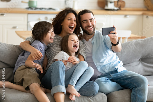 Close up happy parents with little children using smartphone together sitting on couch at home. Smiling father holding phone taking selfie with kids. Family watching video having fun with cellphone. © fizkes
