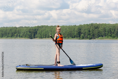 Sup surfing on the lake. Beautiful girl stands on a surfboard with a paddle in her hand. Orange vest. Summer day.