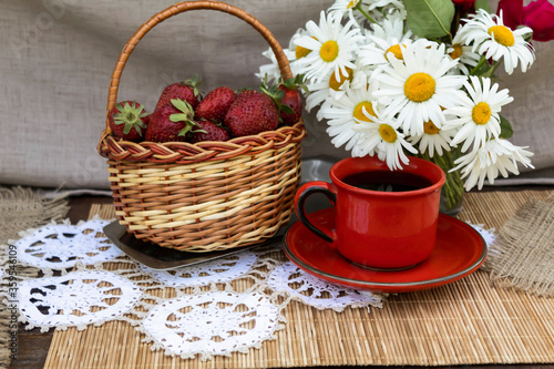On a tracery napkin is a basket with strawberries, a bouquet of daisies and a red cup of coffee.