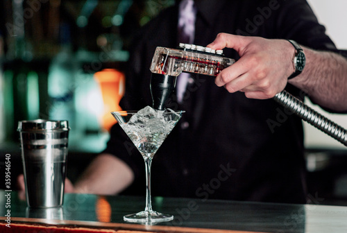 Bartender uses fountain soda gun to finish the martini style wine glass filled with ice, near a shaker at a bar counter.