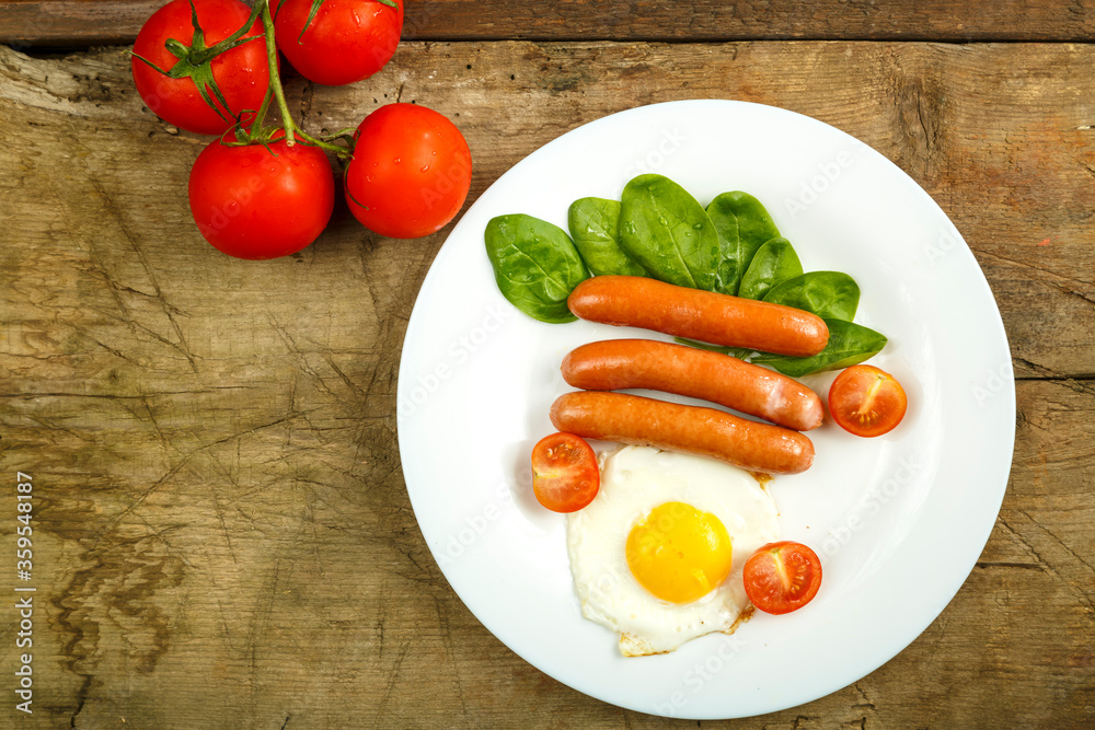 White plate with fried egg with sausages and tomatoes on a wooden table. White plate with fried egg with sausages and tomatoes on a wooden table.