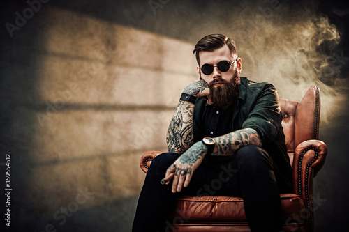 Cool bearded young man sitting on a vintage chair in a dark studio with smoke and window silhouette in the background