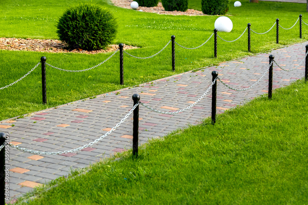 stone walkway and tiles in the park with a fence made of black columns and a chain stretched between the supports in the garden with green grass of the backyard close up nobody.