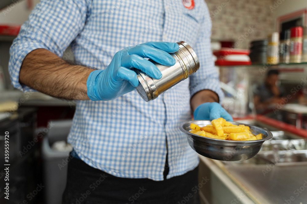 Close up on hands midsection of unknown man entrepreneur with blue rubber protective glove salting potato chips french fries in metal plate at the fast food restaurant in day serving Close up on hands midsection of unknown man entrepreneur with blue rubber protective glove salting potato chips french fries in metal plate at the fast food restaurant in day serving