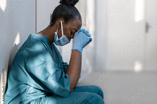 Tired depressed female african scrub nurse wears face mask blue uniform gloves sits on hospital floor. Exhausted sad black doctor feels burnout stress of corona virus frontline protection pray at work photo