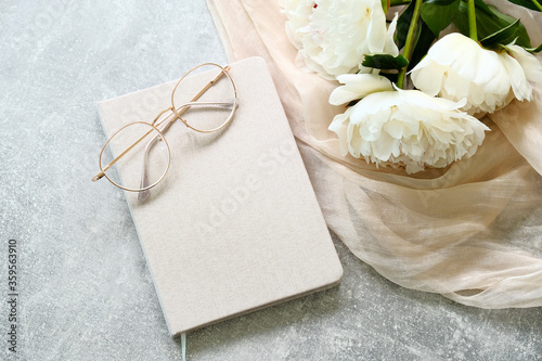Fototapeta Naklejka Na Ścianę i Meble -  Feminine workspace with paper notebook, eyeglasses, peony flowers on stone table. Flat lay, top view. Stylish home office desk.
