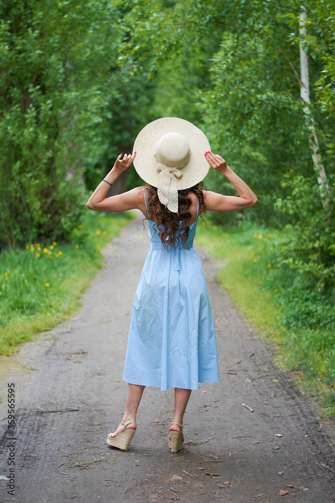 beautiful girl in a straw hat and blue dress on the road in the Park.curly brunette stands with her back to the camera
