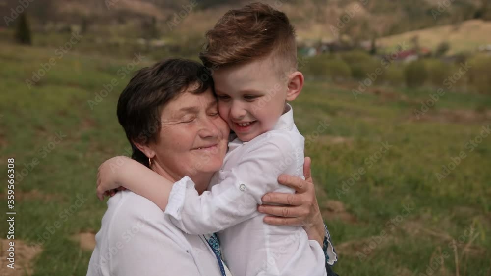 Grandson approaches the grandmother. He puts his arm around her neck. I love my gran. Relax outside on green grass, behind mountain. Bright sunny day Grandson approaches the grandmother. He puts his arm around her neck. I love my gran. Relax outside on green grass, behind mountain. Bright sunny day