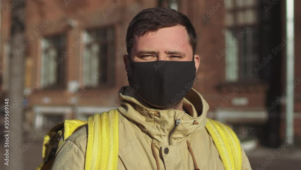 Caucasian male delivery man in a black mask and a jacket with a yellow backpack turns to the camera, takes off his mask and takes a deep breath. Caucasian male delivery man in a black mask and a jacket with a yellow backpack turns to the camera, takes off his mask and takes a deep breath.