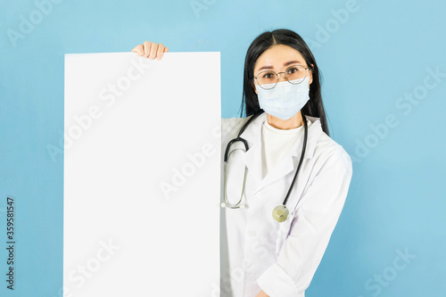 Smart young asian female doctor in lab coat with Medical face mask,white latex medical gloves and stethoscope,carrying a white charging plate against blue background,health care concept