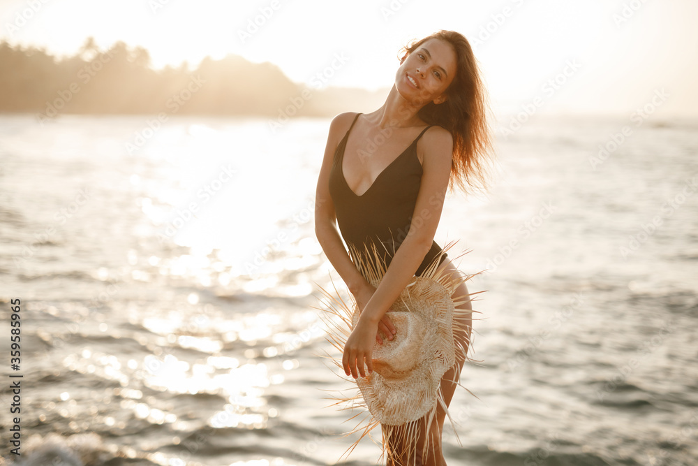 Naklejka premium Portrait of attractive blonde girl woman with long hair posing on rocky beach on sunset background, hold straw hat in her hands and look at the camera