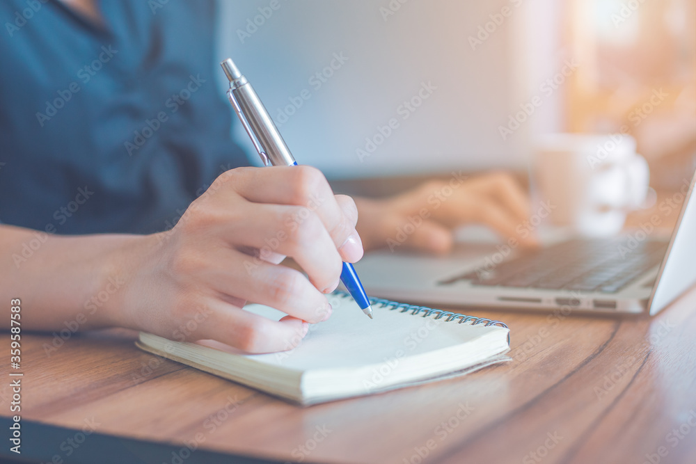 Woman is writing on a notebook with a pen and using a laptop to work in the office.