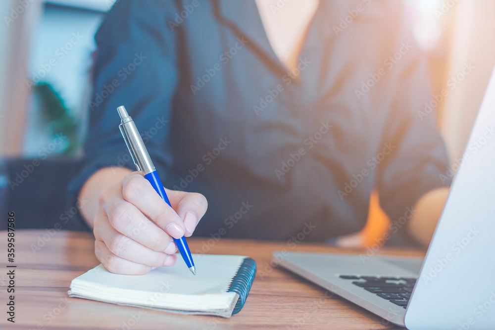 Woman is writing on a notebook with a pen and using a laptop to work in the office.