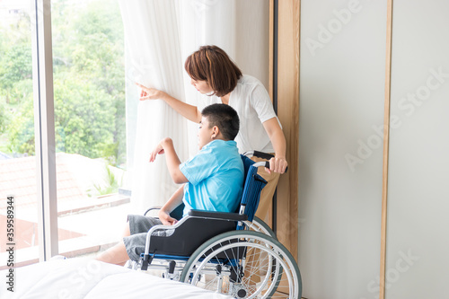asian woman taking care disabled child on wheelchair in bedroom. both looking at something beside of window.