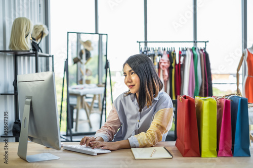 Young Asian woman business owner sitting at a desk in a clothing fashion shop working on a computer next to many color shopping bags.