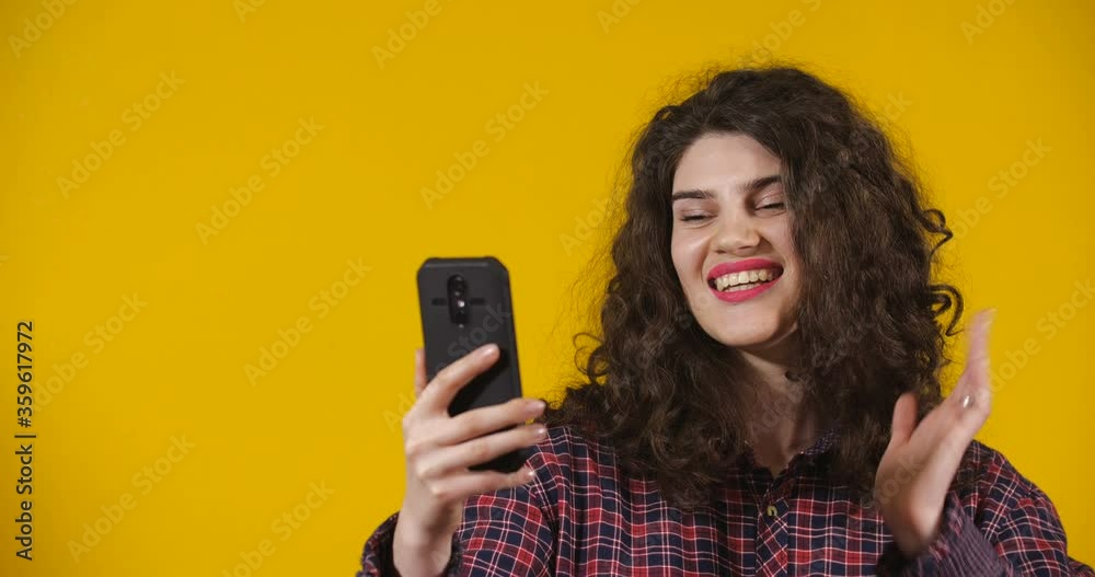 Close-up attractive plus-size model posing. Curly-haired brunette on bright yellow studio background demonstrating different emotions. Friendly girl communicates via video call using smartphone Close-up attractive plus-size model posing. Curly-haired brunette on bright yellow studio background demonstrating different emotions. Friendly girl communicates via video call using smartphone