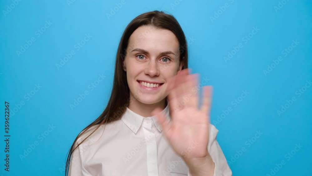 Smiling young brunette woman looking at camera with charming smile waving greeting with hand as notices someone, dressed in white shirt isolated on blue studio background. People lifestyle concept. Smiling young brunette woman looking at camera with charming smile waving greeting with hand as notices someone, dressed in white shirt isolated on blue studio background. People lifestyle concept.