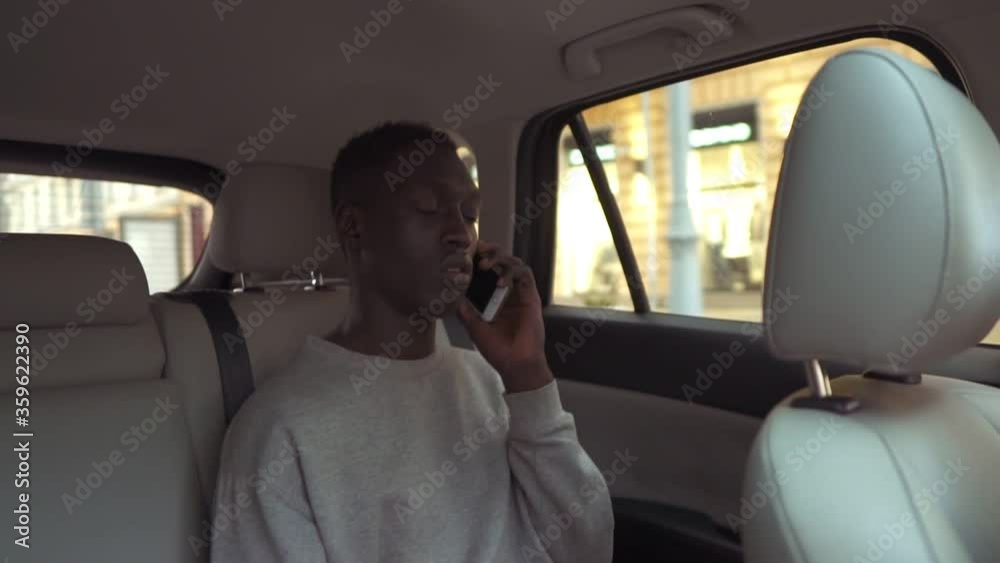 Calm handsome african man in casual clothes talking on cellphone while riding in backseat of taxi cab. Cheerful black passenger guy answering call phone during road trip in the city