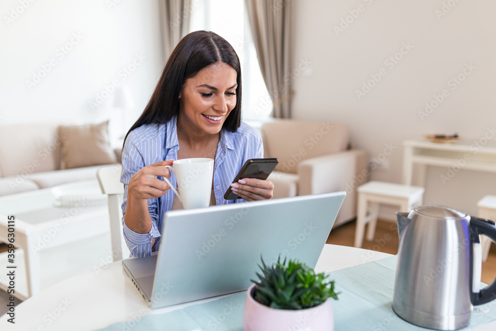 Self employed woman working with her phone and laptop at home with a cup of coffee. Happy woman using mobile phone while working at home with laptop. Smiling woman messaging with smartphone. Self employed woman working with her phone and laptop at home with a cup of coffee. Happy woman using mobile phone while working at home with laptop. Smiling woman messaging with smartphone.
