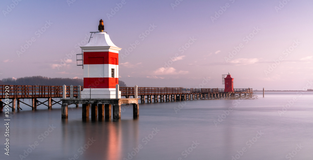 Long exposure shot of the Red Lighthouse of Lignano Sabbiadoro during a cold wintry sunrise. Calm water under a wooden boardwalk. Concept for relax and vacation Long exposure shot of the Red Lighthouse of Lignano Sabbiadoro during a cold wintry sunrise. Calm water under a wooden boardwalk. Concept for relax and vacation