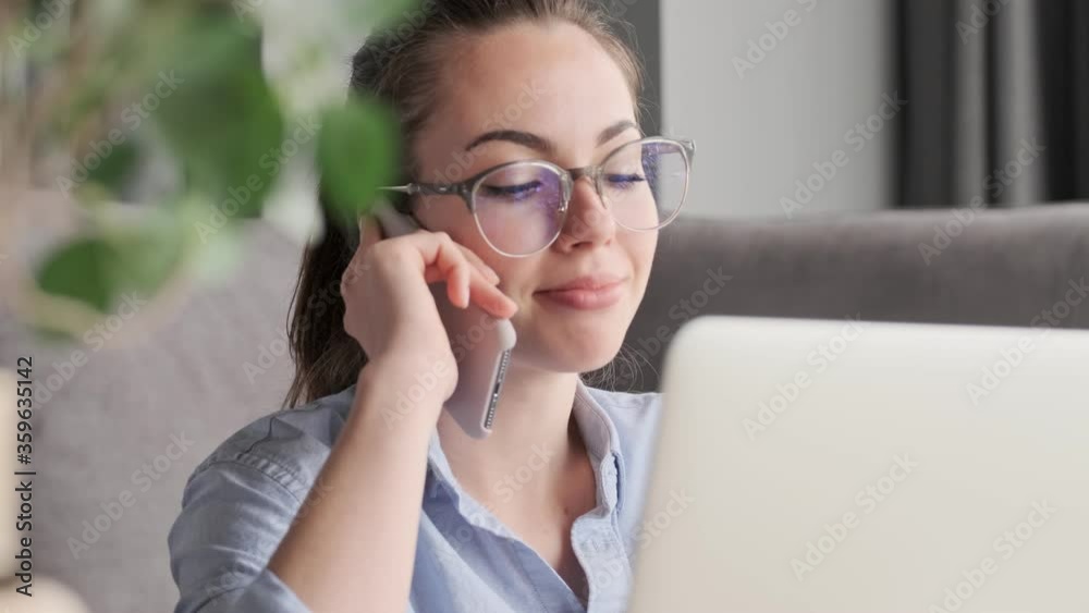 Smiling attractive brunette woman in eyeglasses talking by smartphone while sitting with laptop computer on the floor near sofa at home Smiling attractive brunette woman in eyeglasses talking by smartphone while sitting with laptop computer on the floor near sofa at home