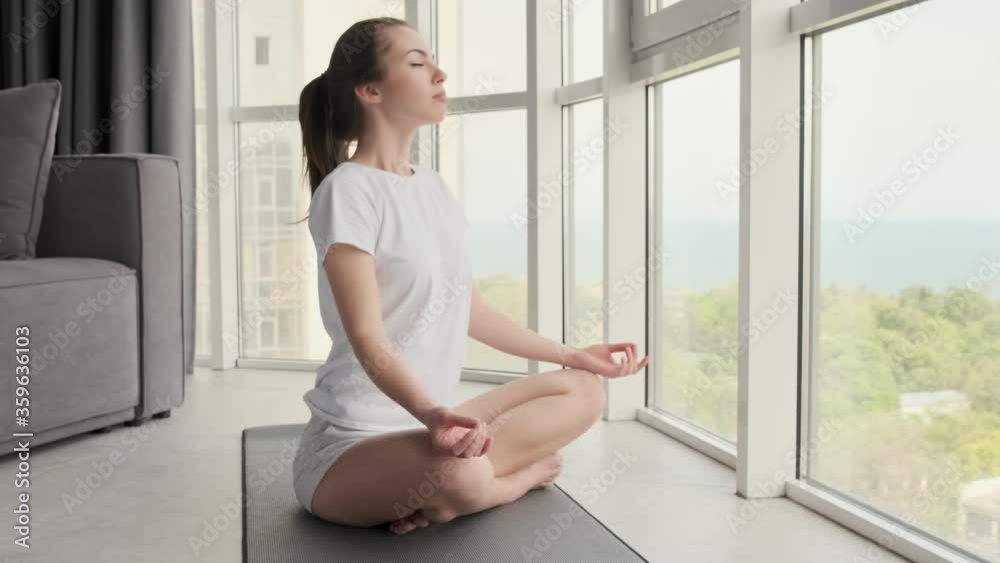 Side view of Carefree attractive brunette woman meditating with closed eyes on the floor near window at home Side view of Carefree attractive brunette woman meditating with closed eyes on the floor near window at home