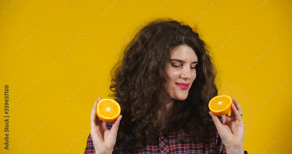 Close-up attractive plus-size model posing. A curly-haired brunette on a bright yellow studio background demonstrating different situations. Smiling Girl closing her eyes with halves of an orange Close-up attractive plus-size model posing. A curly-haired brunette on a bright yellow studio background demonstrating different situations. Smiling Girl closing her eyes with halves of an orange