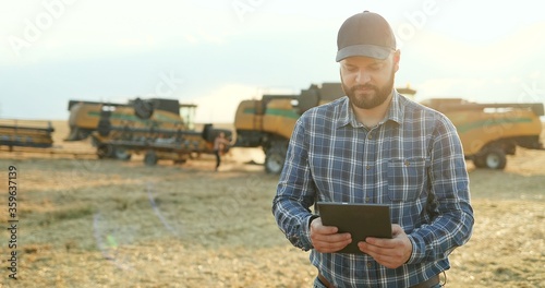 Caucasian man in a hat standing in the field and calculating amount of grain while noting in the documents while machines gathering harvest behind him.