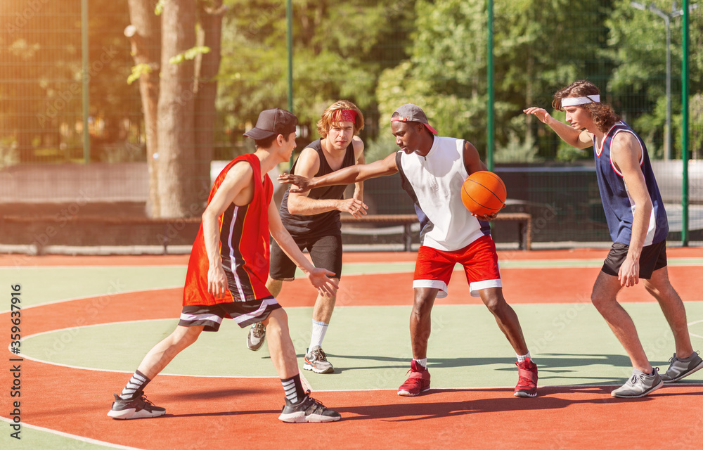 Team of millennial sportsmen engaged in basketball game at outdoor court in summer