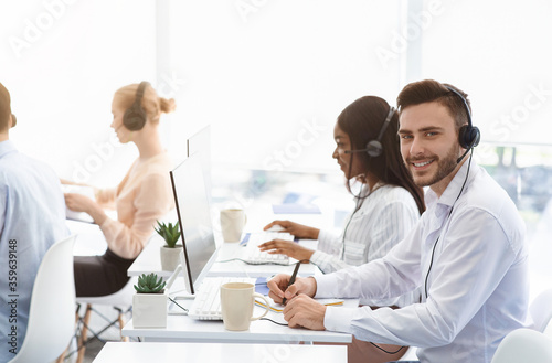 Attractive call center operator smiling at camera and his coworkers talking to clients at office, copy space