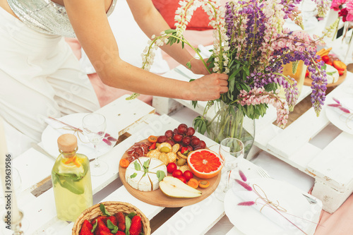 Cut view of young florist woman decorating with beautiful lupine flowers a festive summer picnic for wedding or birthday celebration in light pink colors in the garden. Food and drink concept