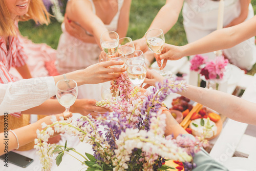 Group of young women have fun at summer picnic  in the garden with tasty food and white wine. Cheers with wineglasses. Birthday celebration or hen-party. Friendship. Flowers decoration